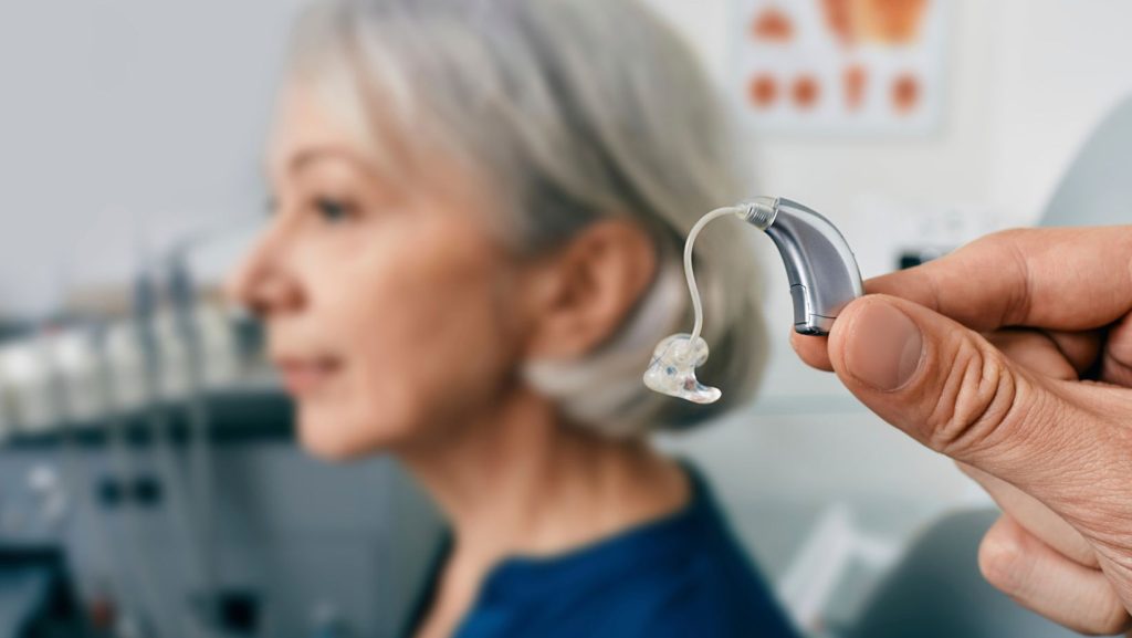 Close up of two fingers holding a single hearing aid with an older woman in the background.