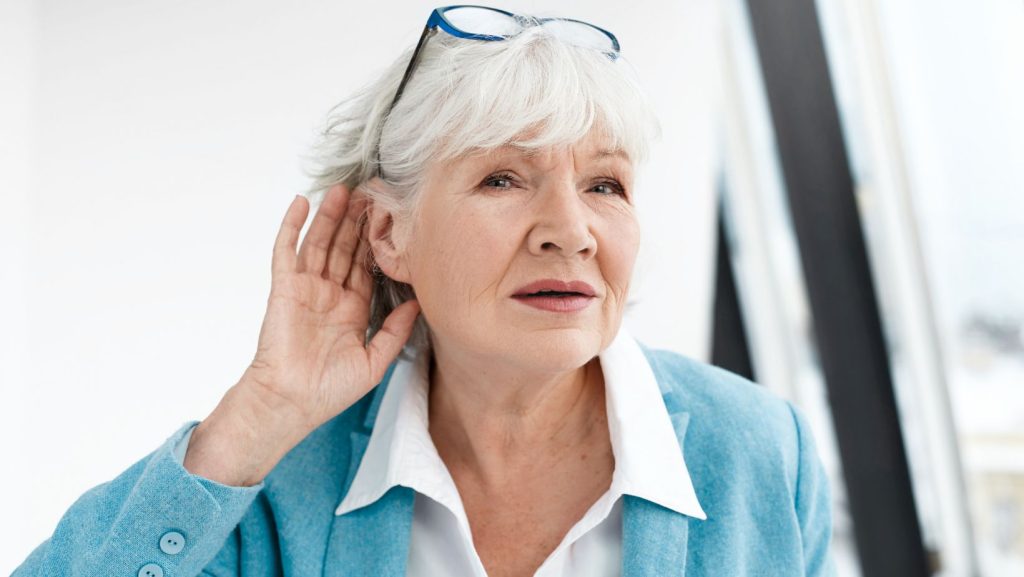 Elderly woman with short white hair, wearing a light blue jacket, leans forward with a hand to her ear, struggling with hearing loss.